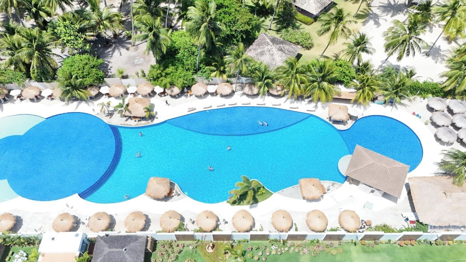 An aerial view of a resort with a large swimming pool, palm trees, and sun loungers.