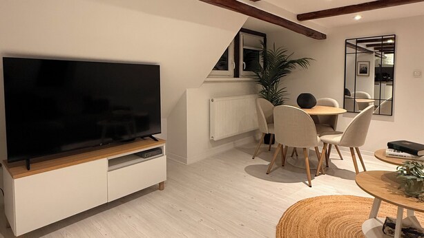 En moderne stue med et stort fladskærms-TV, træbjælker i loftet, et spiseområde med et rundt bord og en radiator.