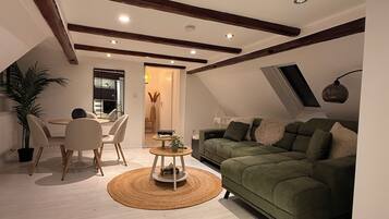 Ein gemütlicher Lofts-Wohnraum mit hoher Decke, freiliegenden Holzbalken, einem grünen Sofa, einer Essecke mit rundem Tisch und einer Topfpflanze.