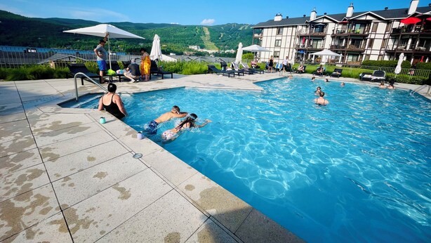 Una piscina con gente, un suelo de baldosas y vistas a colinas verdes al fondo.