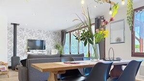 A modern living room with a sofa, a wooden dining table, and chairs. There is a TV on the wall and a plant on the windowsill. - Gîte marais Audomarois, pedestrian/boat access (Tilques)