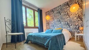 A bedroom with a bed, a small table, a chair, a window with curtains, and a wall with a leaf pattern. - Gîte marais Audomarois, pedestrian/boat access (Tilques)