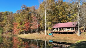 A cabin with a red roof, surrounded by autumn-colored trees and a calm lake.