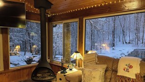 A cozy cabin interior with a fireplace, large windows with snowy forest views, a comfortable sofa, and a small table with a lamp.