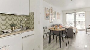 Eine moderne Wohnung mit Küche, Essbereich und Wohnzimmer. Die Küche verfügt über weiße Schränke und eine geflieste Spritzschutzleiste. Der Essbereich bietet einen runden Tisch mit Stühlen. Das Wohnzimmer ist mit einer Couch und einem großen Fenster ausgestattet.