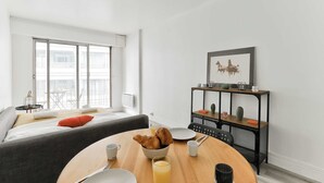 Ein modernes Wohnzimmer mit einer Couch, einem Esstisch mit Frühstücksgebäck und -zutaten, einem Fenster mit Aussicht und einem dekorativen Regal.