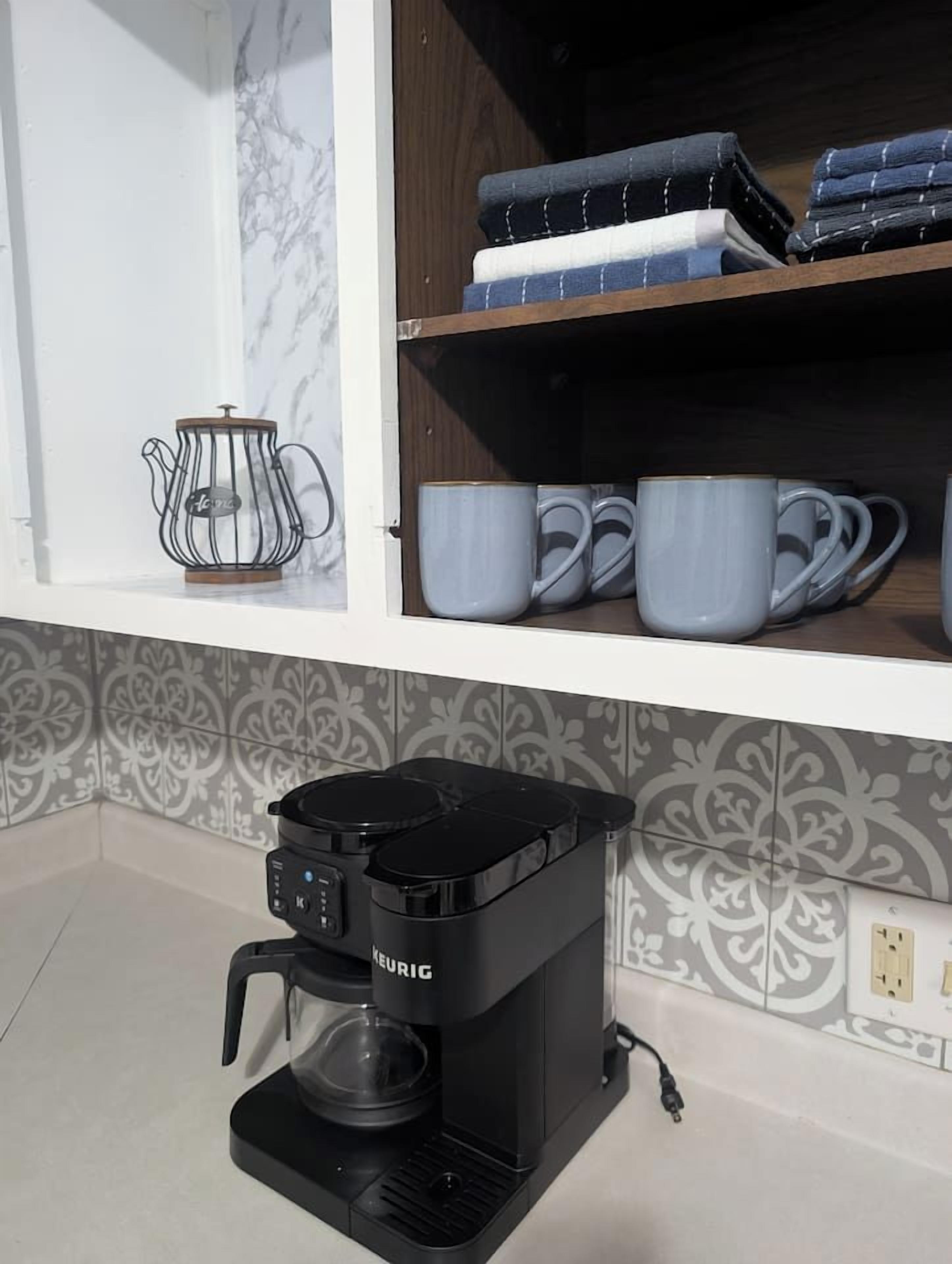 Une cuisine équipée d’une machine à café Keurig, d’une bouilloire et d’une étagère où sont rangées des serviettes pliées et des tasses.