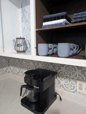 A kitchen with a Keurig coffee maker, a kettle, and a shelf with folded towels and mugs.