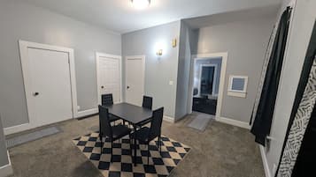 Une pièce avec un tapis à carreaux, une table entourée de chaises et une porte donnant sur une autre pièce.
