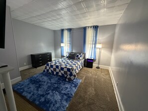 Ein Schlafzimmer mit karierter Bettdecke, einem Schminktisch, einer Lampe und einem Fenster mit Vorhängen.