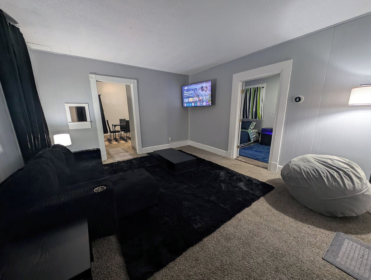 Un salon avec un canapé noir, un fauteuil poire, un téléviseur à écran plat fixé au mur et une porte-fenêtre donnant sur une autre pièce.