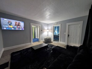 A living room with a flat-screen TV mounted on the wall, a black sofa, a small coffee table, and a door leading to a bedroom. - Work Crews: Clean 2BR near US-41 + Laundry & TVs (Evansville)