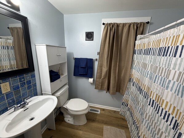 Ein Badezimmer mit einem weißen Waschbecken, einer Toilette und einem Schrank. Ein blaues Handtuch hängt an der Wand und ein beiges Duschvorhang ist angebracht.