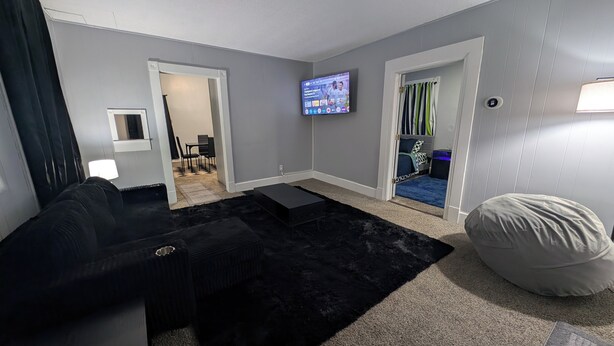 Ein Wohnzimmer mit schwarzem Sofa, schwarzem Teppich, Fernseher an der Wand und einem Bett mit gestreiftem Vorhang.