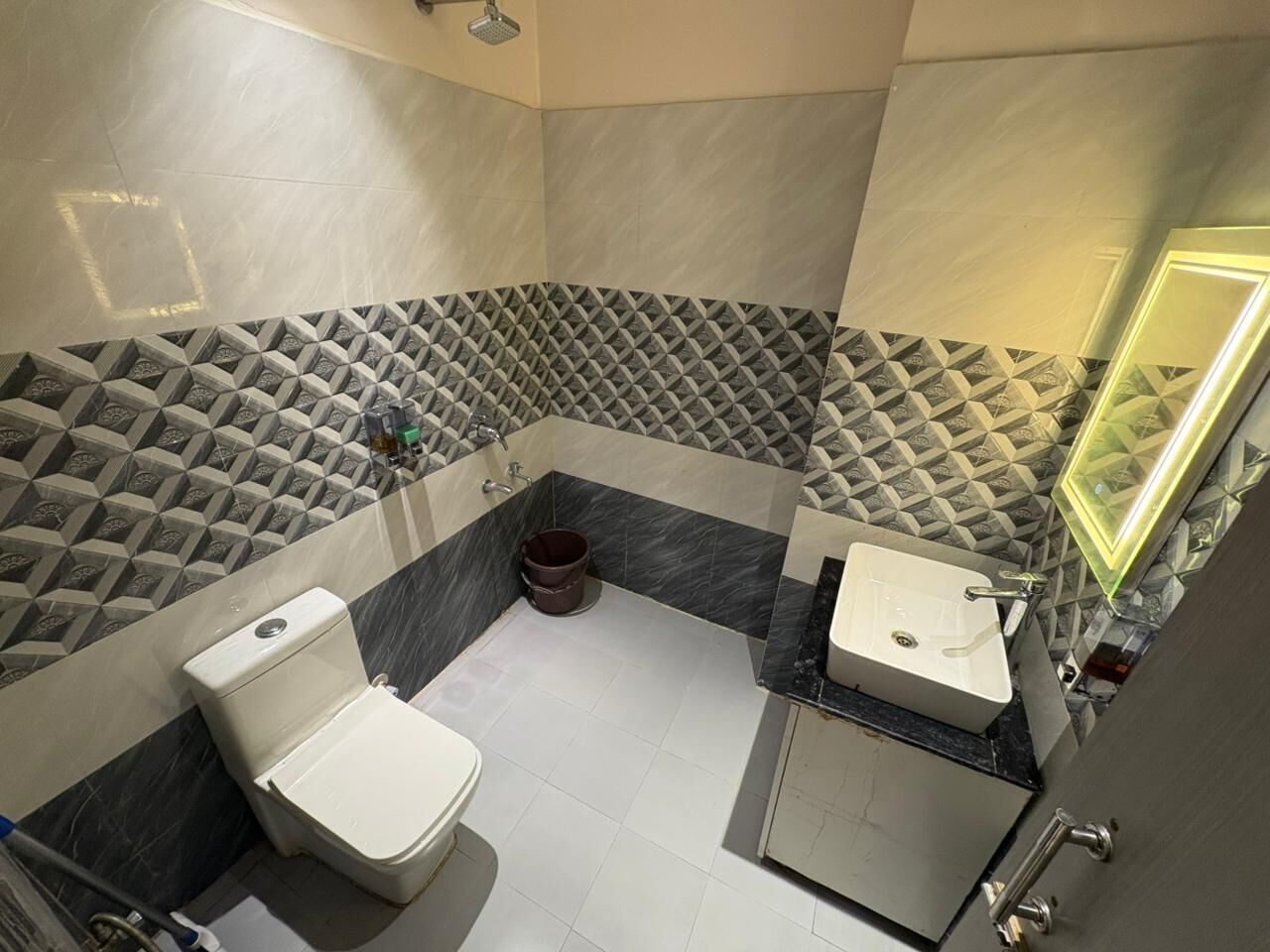 Et badeværelse med toilet, bidet og vask, med geometriske fliser og et spejl med lys.