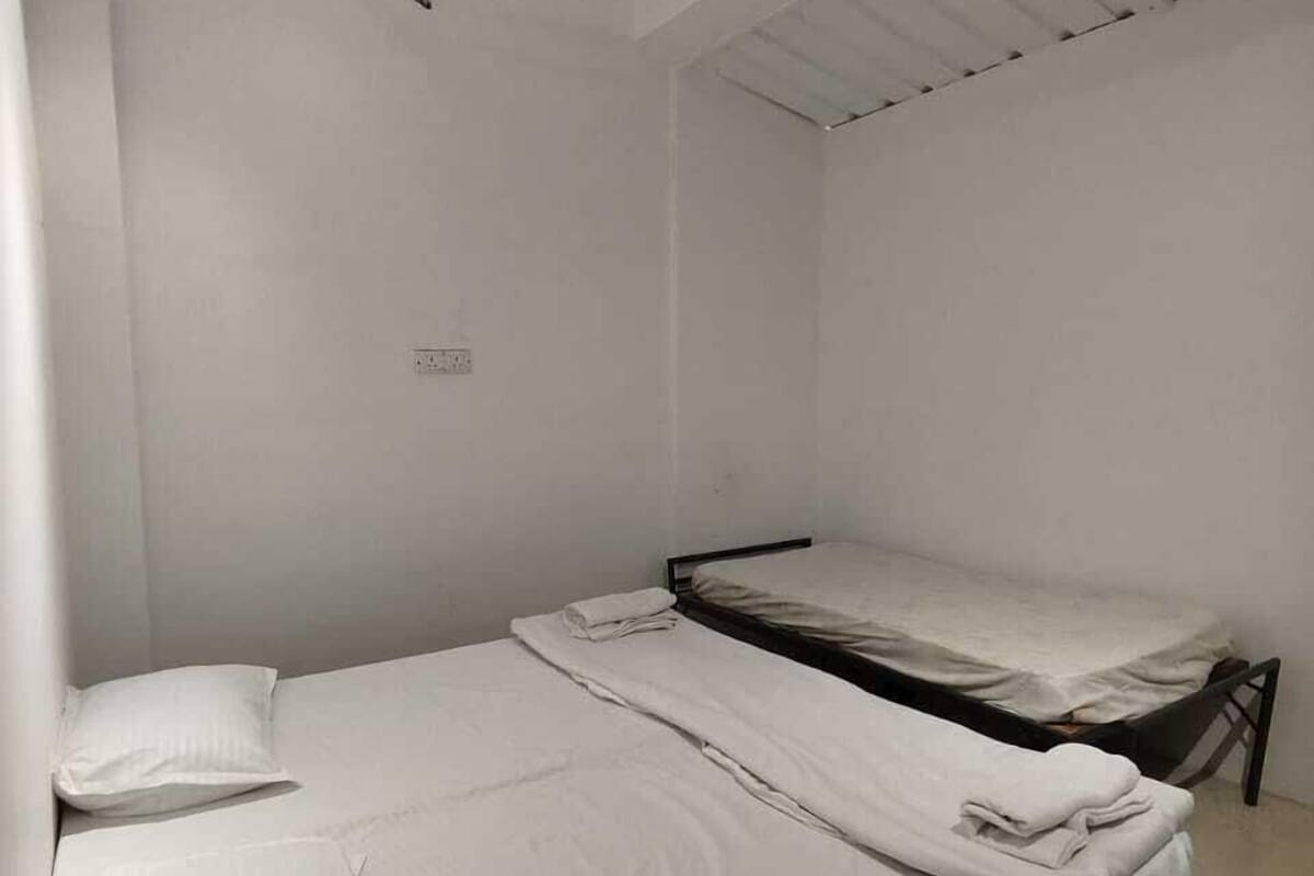 A small, single-room hotel room with a bed, a ceiling fan, and a light fixture.