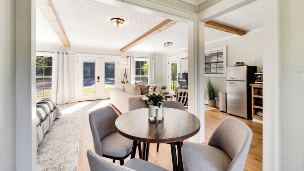 A spacious living area with a round wooden table, grey upholstered chairs, and a sofa. There is a kitchen area with a refrigerator and a dining table. The room has large windows and a high ceiling with exposed beams. - Cozy Mountain Cottage Near Downtown (Highlands)
