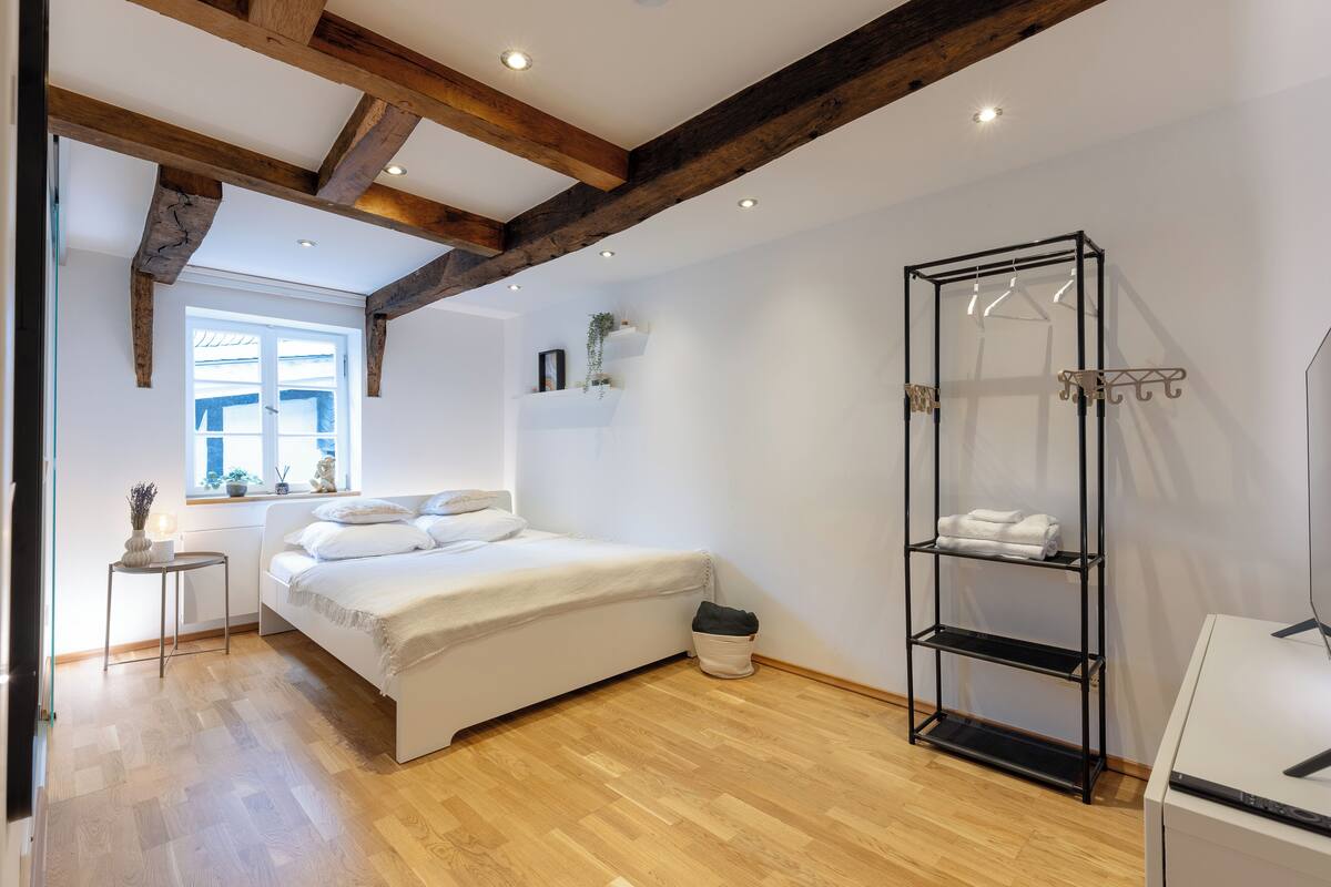 A bedroom with a bed, wooden beams, a window, a bedside table, a hanging shelf, and a radiator.