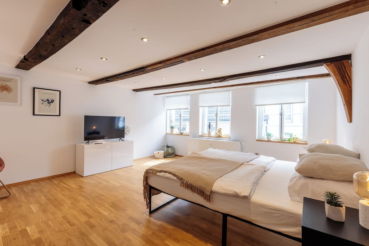 A bedroom with a wooden ceiling, a bed with a beige blanket, a TV on a white cabinet, and windows with plants.