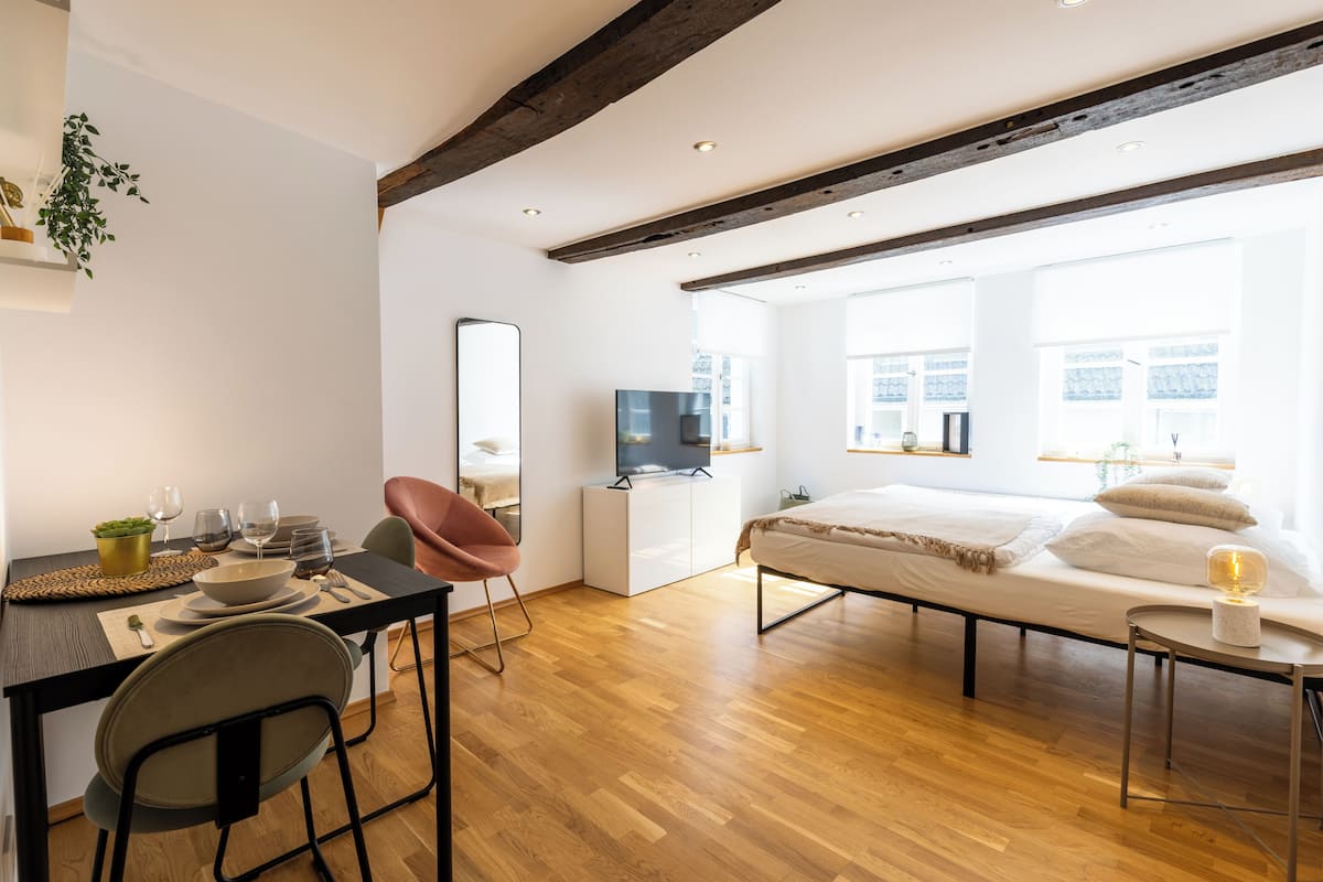 A modern hotel room with a wooden floor, a bed with white linens, a desk with a chair, a television, and large windows offering natural light.