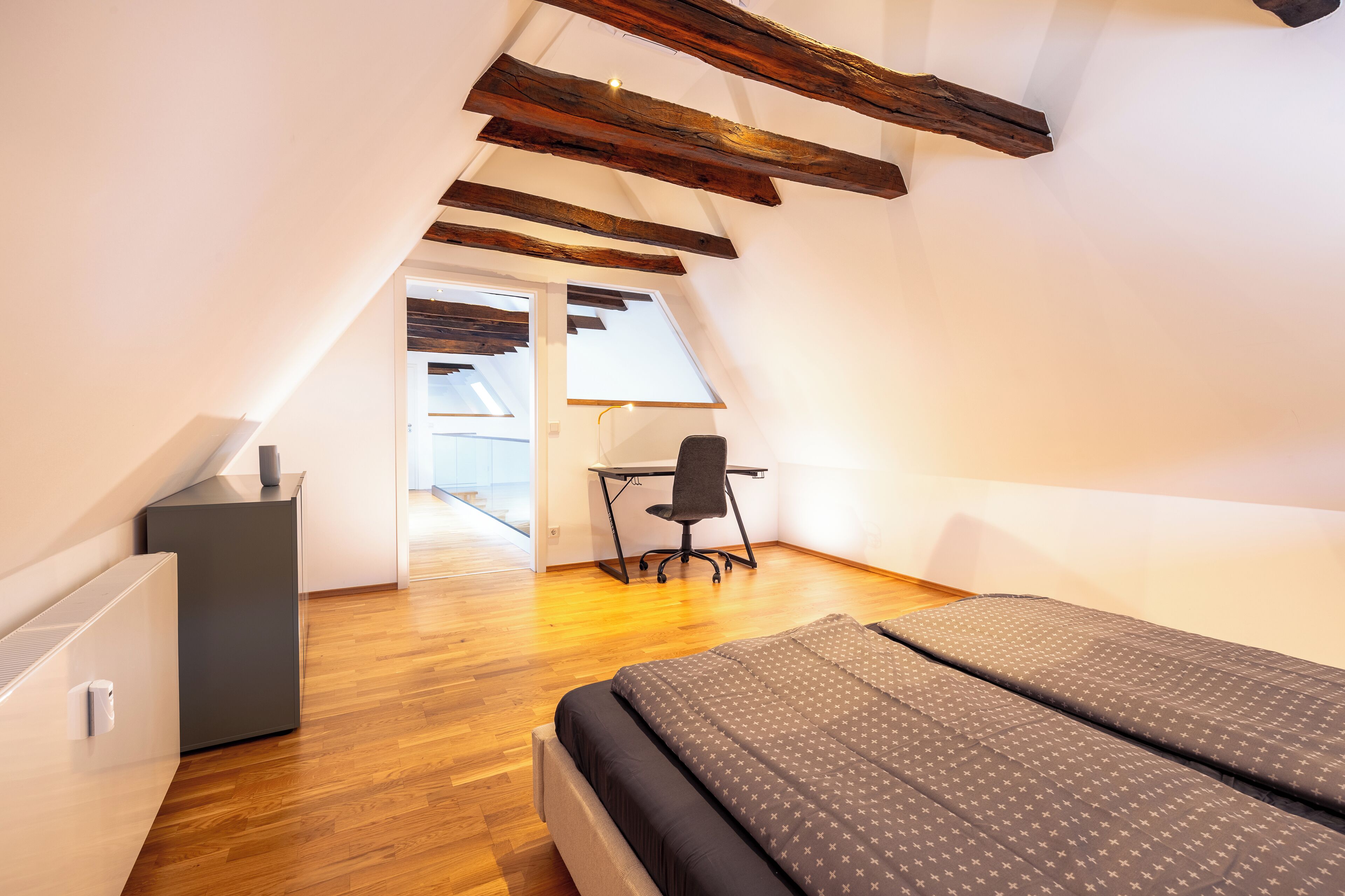 Ein Schlafzimmer mit schrägverdeckter Decke, Holzbalken, einem Bett mit gemusterten Betttextilien, einem Schreibtisch und einem Stuhl.