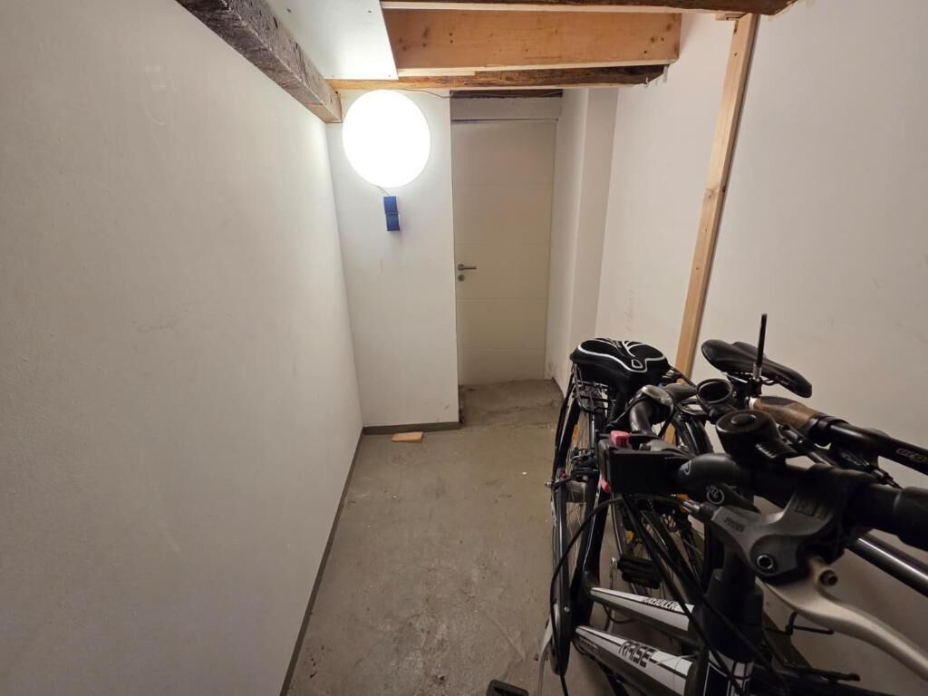 Un couloir étroit, avec un support à vélos sur le côté droit, une porte au fond et un luminaire rond fixé au mur.