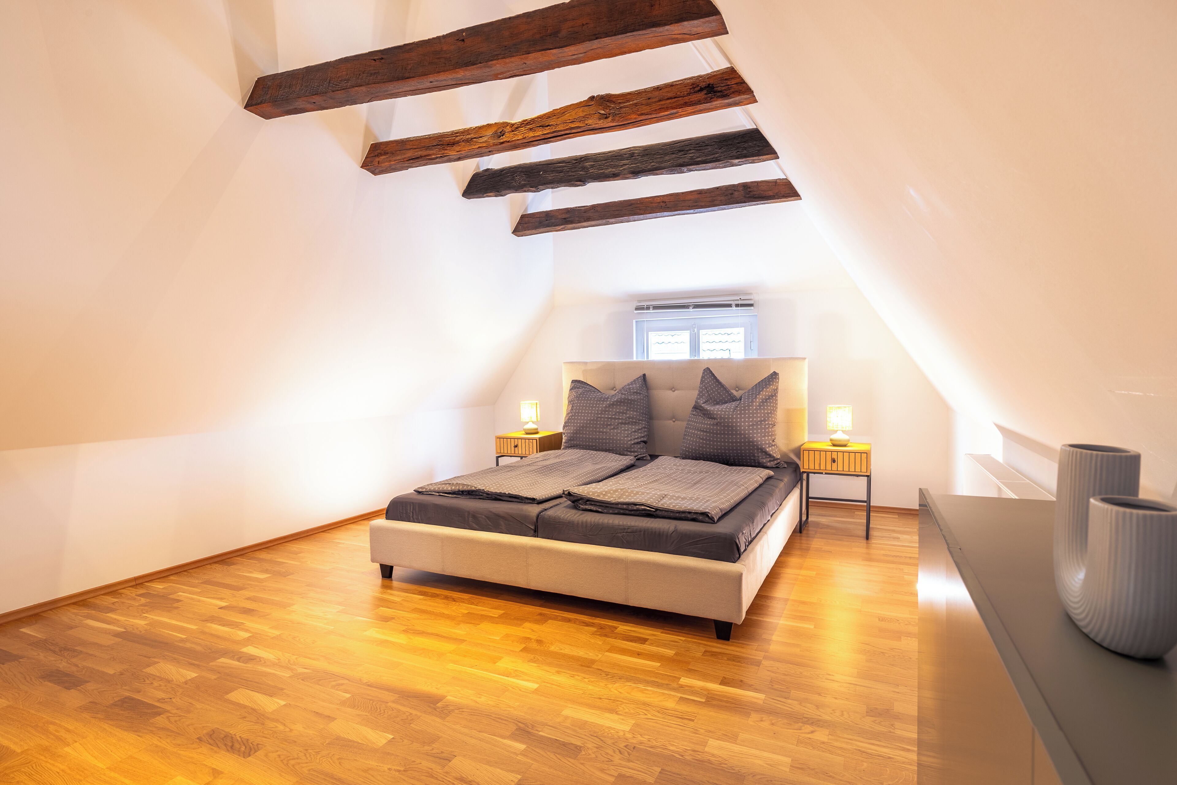 Un dormitorio con techo inclinado, vigas de madera, cama con dos almohadas, lámparas de noche y mesita.