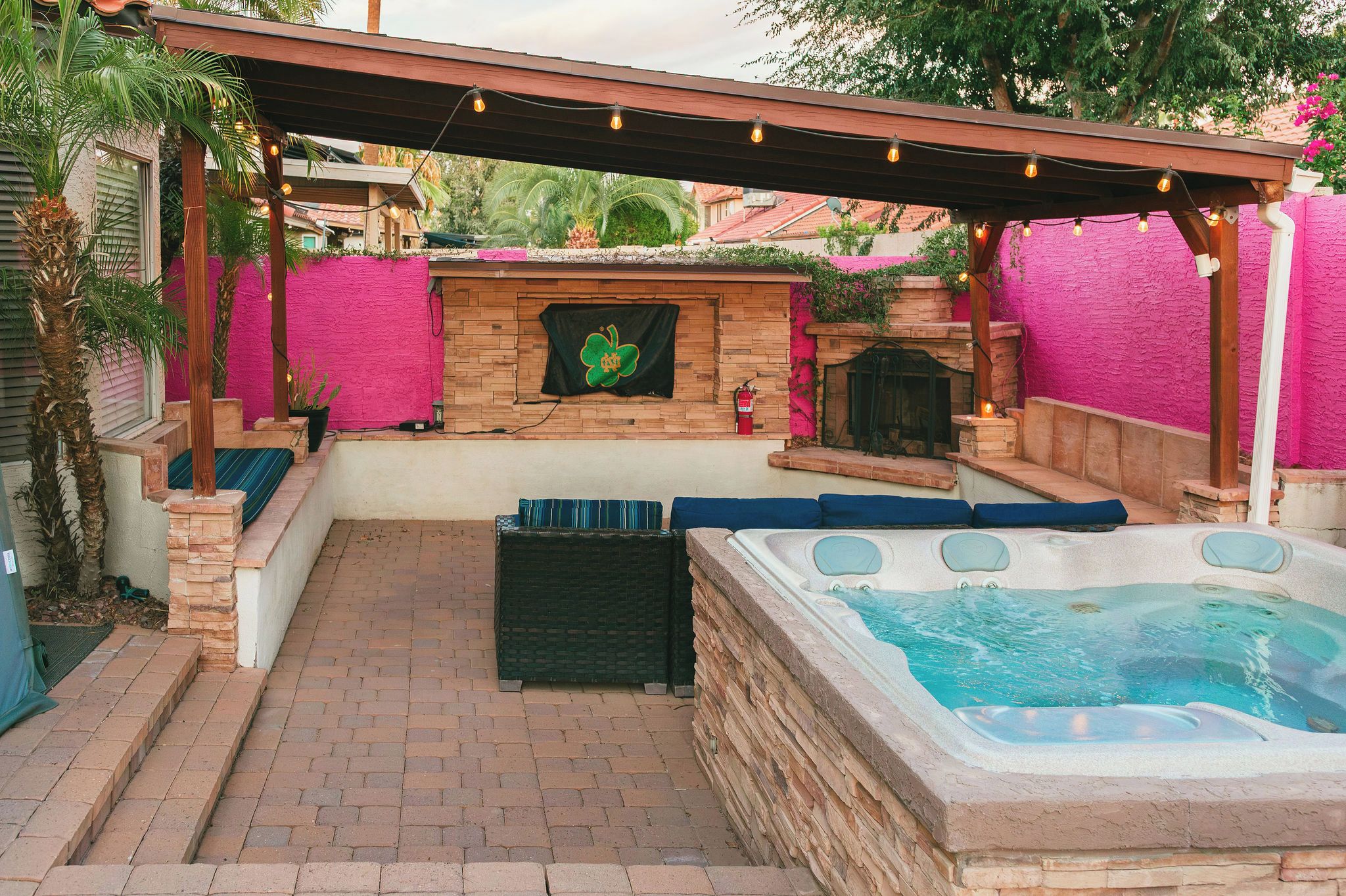 A hot tub area with a stone fireplace, outdoor seating, and a pergola with string lights.