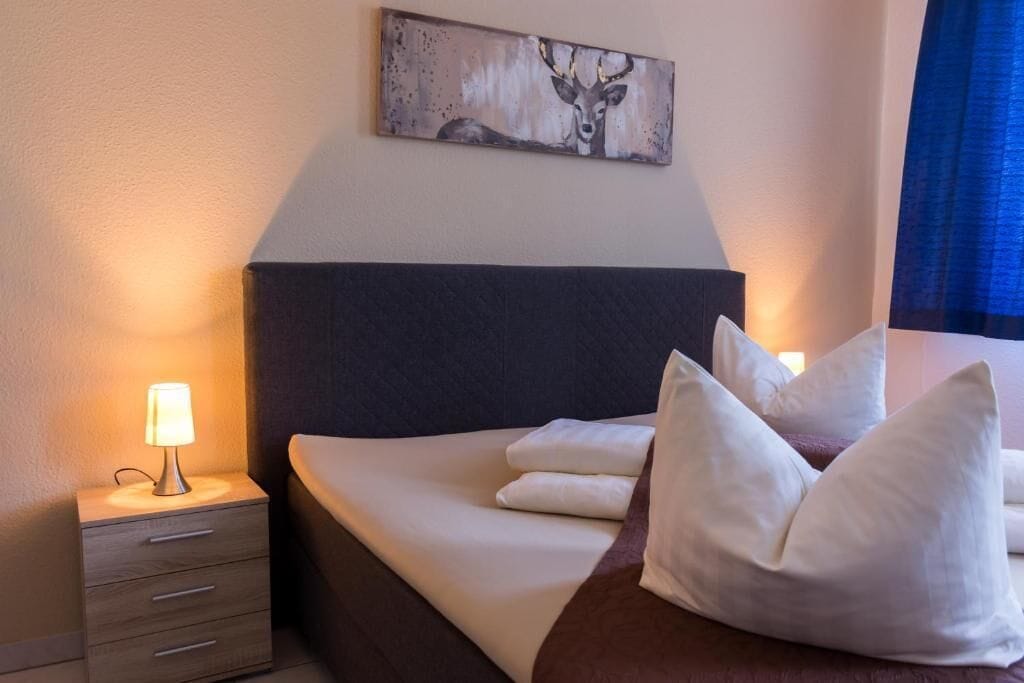 Cama bem arrumada com roupa de cama e almofadas brancas, luminária de cabeceira, mesa de cabeceira de madeira com gavetas e um quadro de um veado pendurado na parede.