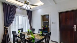 A dining area with a table set for four, surrounded by chairs, a ceiling fan, and a view of a kitchen area. - Luxurious Getaway: 24/7 Security - Pool - Balcony! (Sakumono)