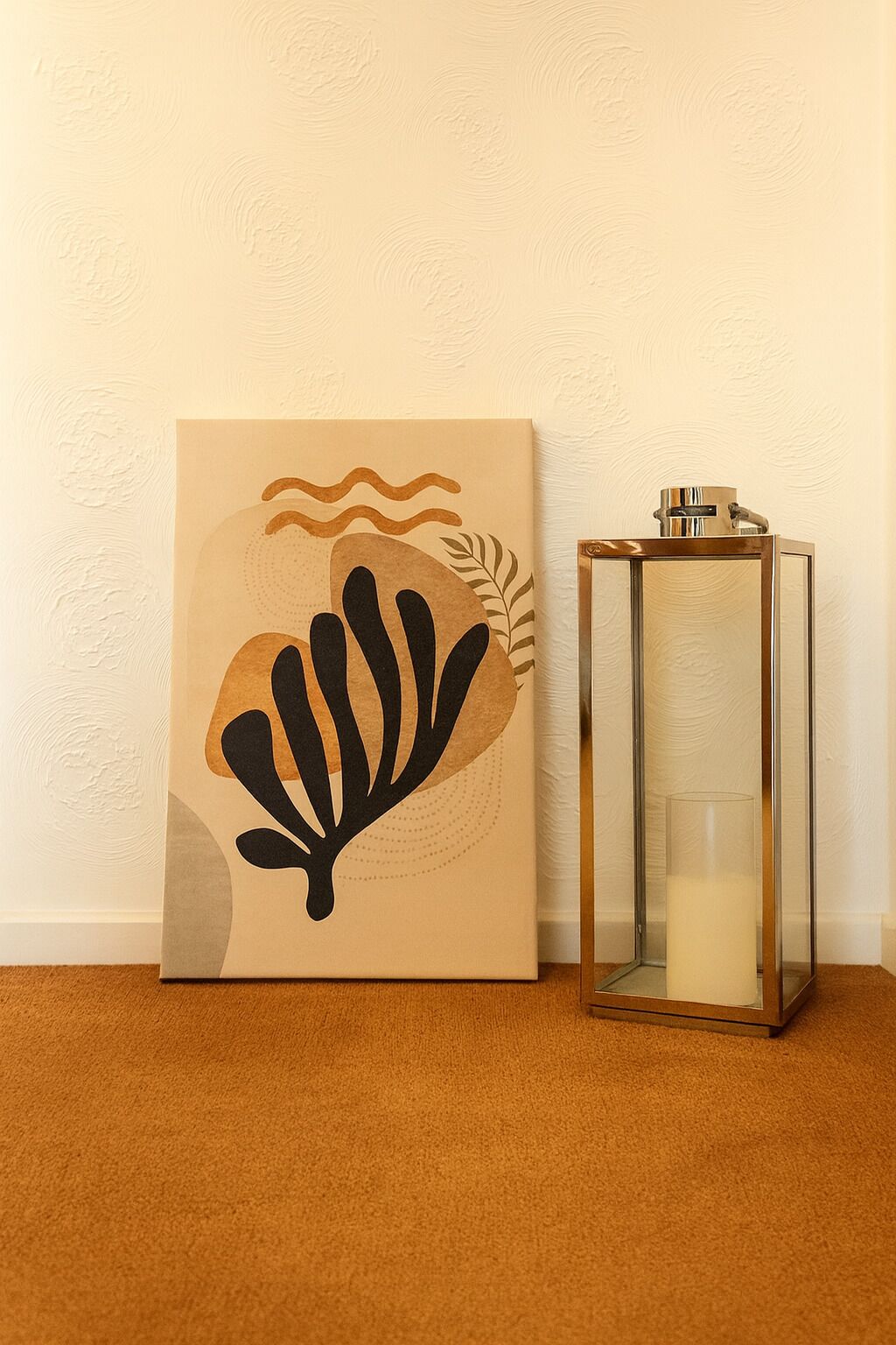 A framed artwork with a leaf design and a glass candle holder on a carpeted floor against a textured wall.