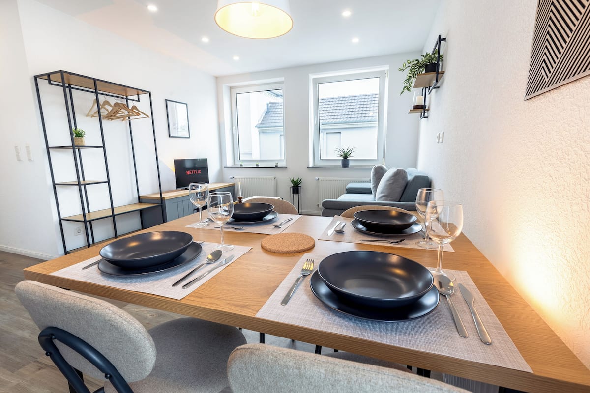 Un comedor moderno con una mesa de madera dispuesta para dos comensales, con platos negros, cubiertos y copas de vino.
