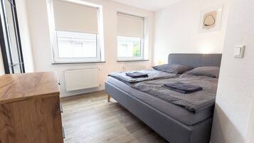 A bedroom with a bed, a wooden dresser, and two windows with blinds.