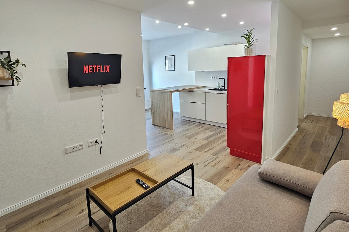 Una sala de estar moderna con un televisor de pantalla plana montado en la pared, una mesa de centro de madera, un sofá y una cocina al fondo.