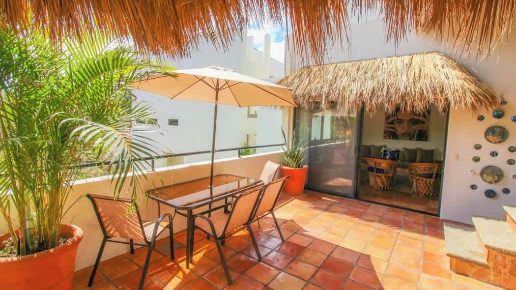 Ein Balkon mit Tisch und Stühlen, Blumentöpfen, einem Strohdach und einer Glastür zu einem Innenraum mit Korbmöbeln.