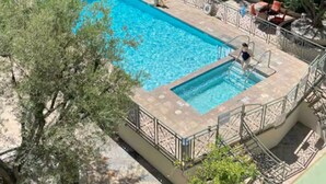 En rektangulær swimmingpool med hegn, omgivet af træer og en bygning med flere etager og balkoner.