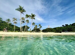 A beach with clear turquoise water, palm trees, and a sandy shore. - Threeos Cabana has simple yet stylish & cozy 1-bedroom unit at Boracay Newcoast. (Aklan)