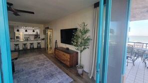 Una sala de estar moderna con una cocina al fondo, un televisor de pantalla plana grande y un balcón con mobiliario exterior.