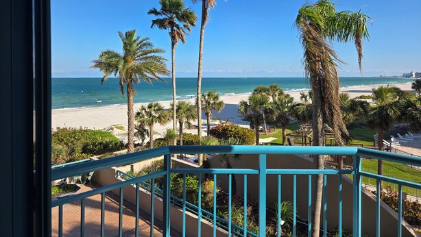 A balcony with a view of a beach, palm trees, and a clear blue sky. - Remodeled 2-bedroom condo in serene Sand Key, Clearwater Beach (Clearwater)