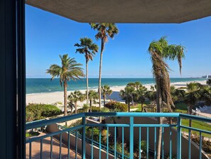 Un balcón con vista a una playa, palmeras y un cielo azul despejado.