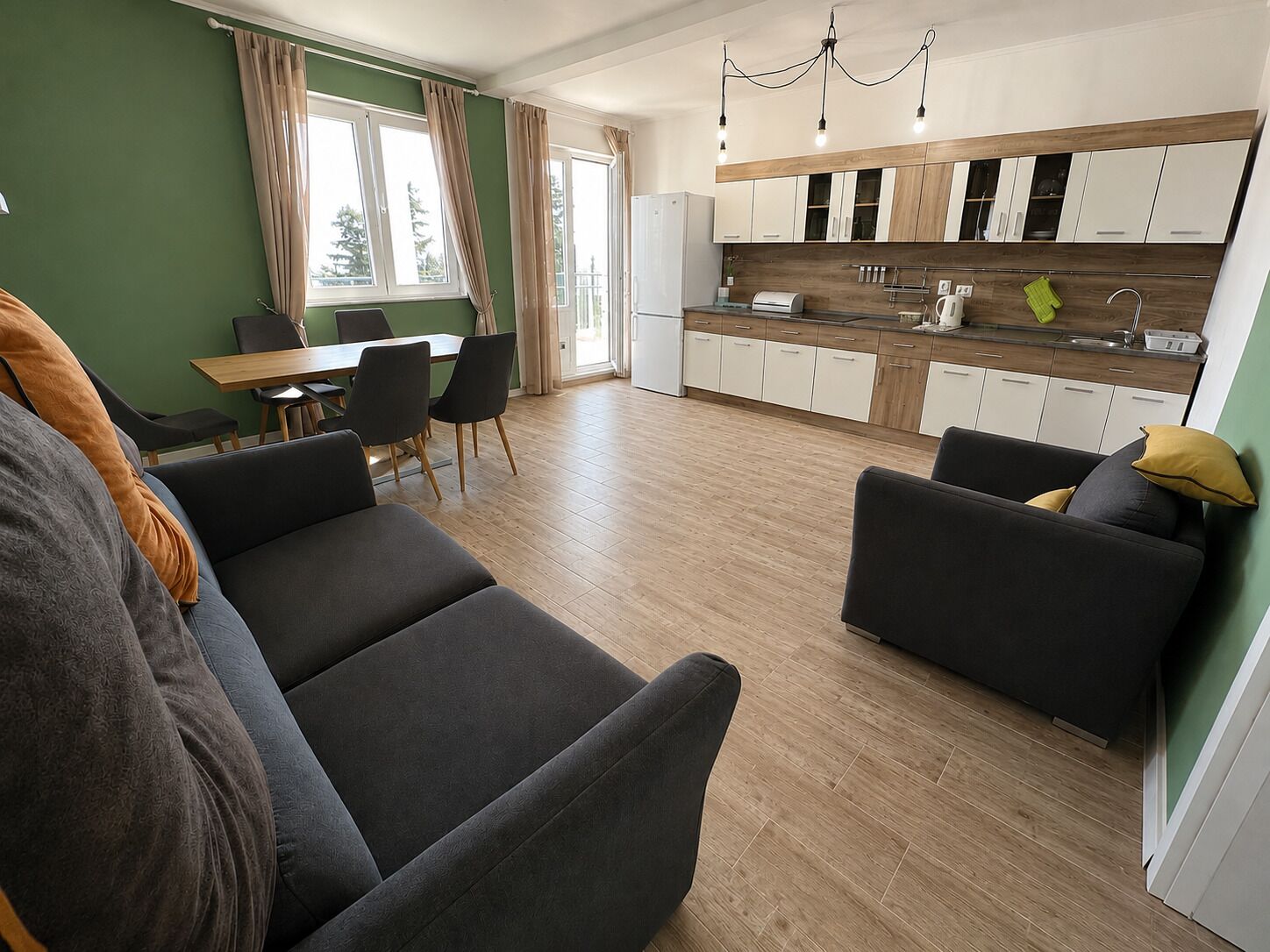 A modern living room with a sofa, a dining table with chairs, a kitchen with wooden cabinets, and a large window with curtains.