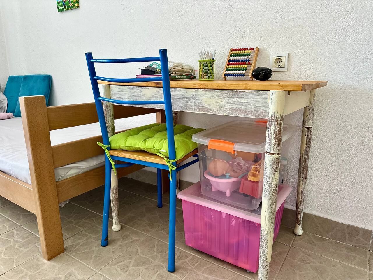 A room with a wooden bed, a blue chair with a green cushion, a small table with a chalkboard, and a pink storage box.