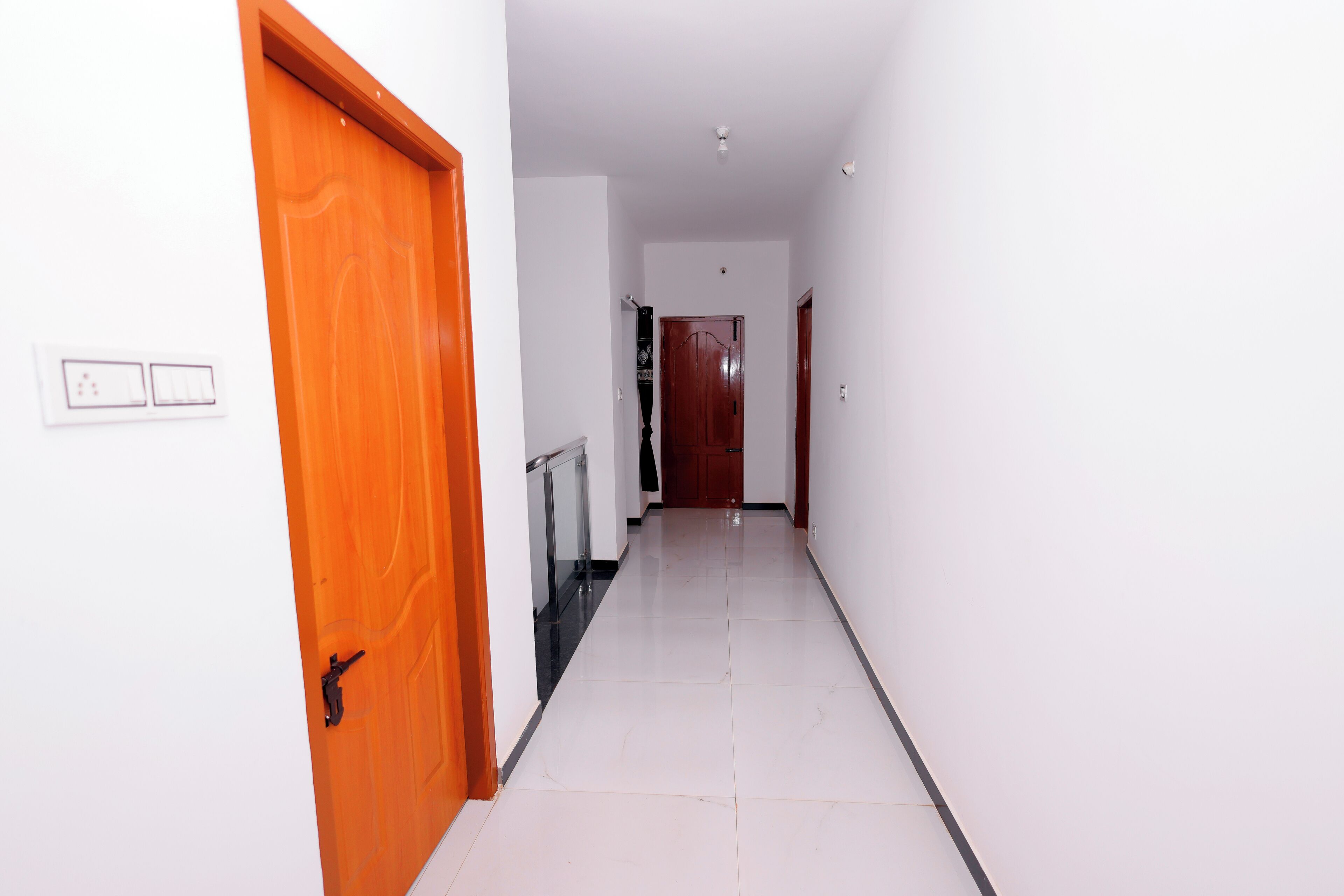 A hallway with a wooden door on the left, a white wall with a light switch, and a tiled floor.