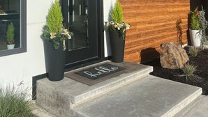 Un edificio moderno con puerta negra, ventanas amplias y fachada de madera. Hay plantas en macetas y un felpudo en el exterior.