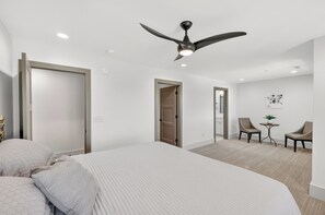Una habitación de hotel con una cama, un ventilador de techo, una pequeña mesa con sillas y una puerta que da a otra habitación.
