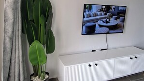 Ein modernes Wohnzimmer mit einem an der Wand befestigten Flachbildfernseher, einem weißen Konsolentisch, einer großen grünen Pflanze und einem grauen Teppich mit Muster.
