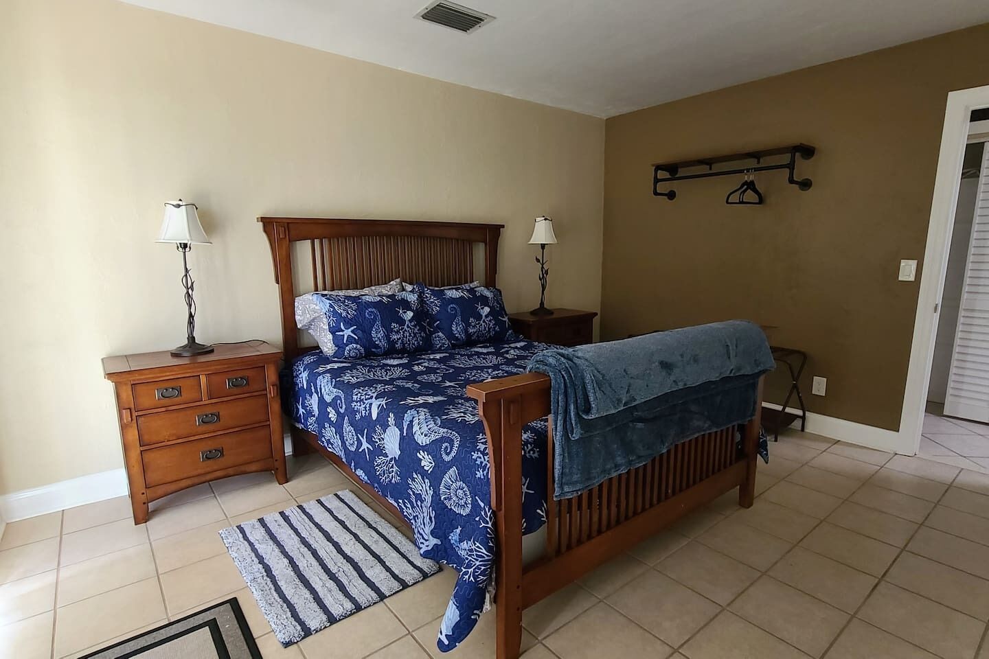 A bedroom with a wooden bed, two bedside tables with lamps, a wall-mounted shelf, and a tiled floor.
