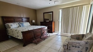 Une chambre avec un grand lit, une commode, une chaise et une fenêtre avec des stores.