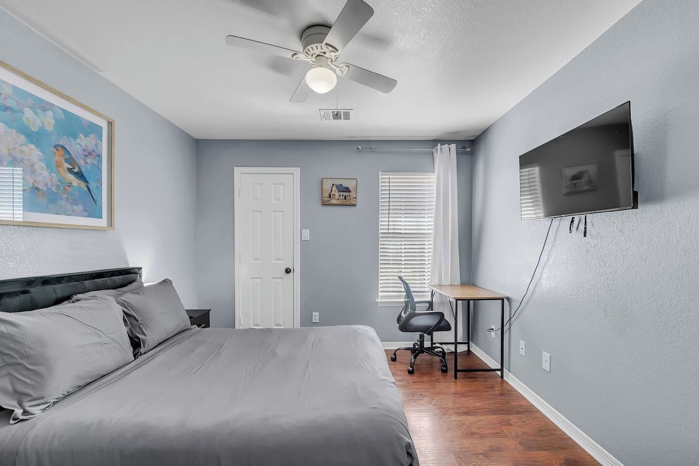 A bedroom with a bed, a desk, a chair, a ceiling fan, a TV, and a door.