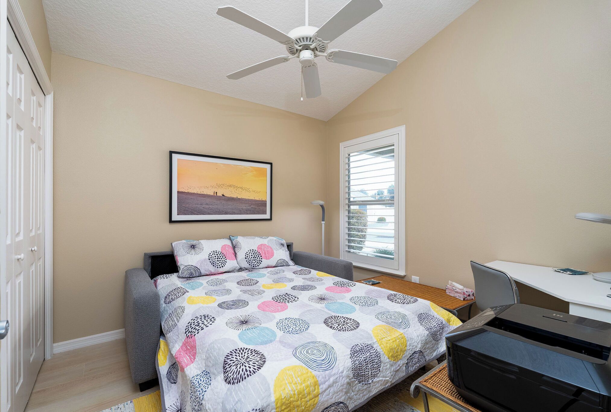 Une chambre à coucher avec un lit, un bureau, un ventilateur de plafond, une fenêtre et un tableau au mur.